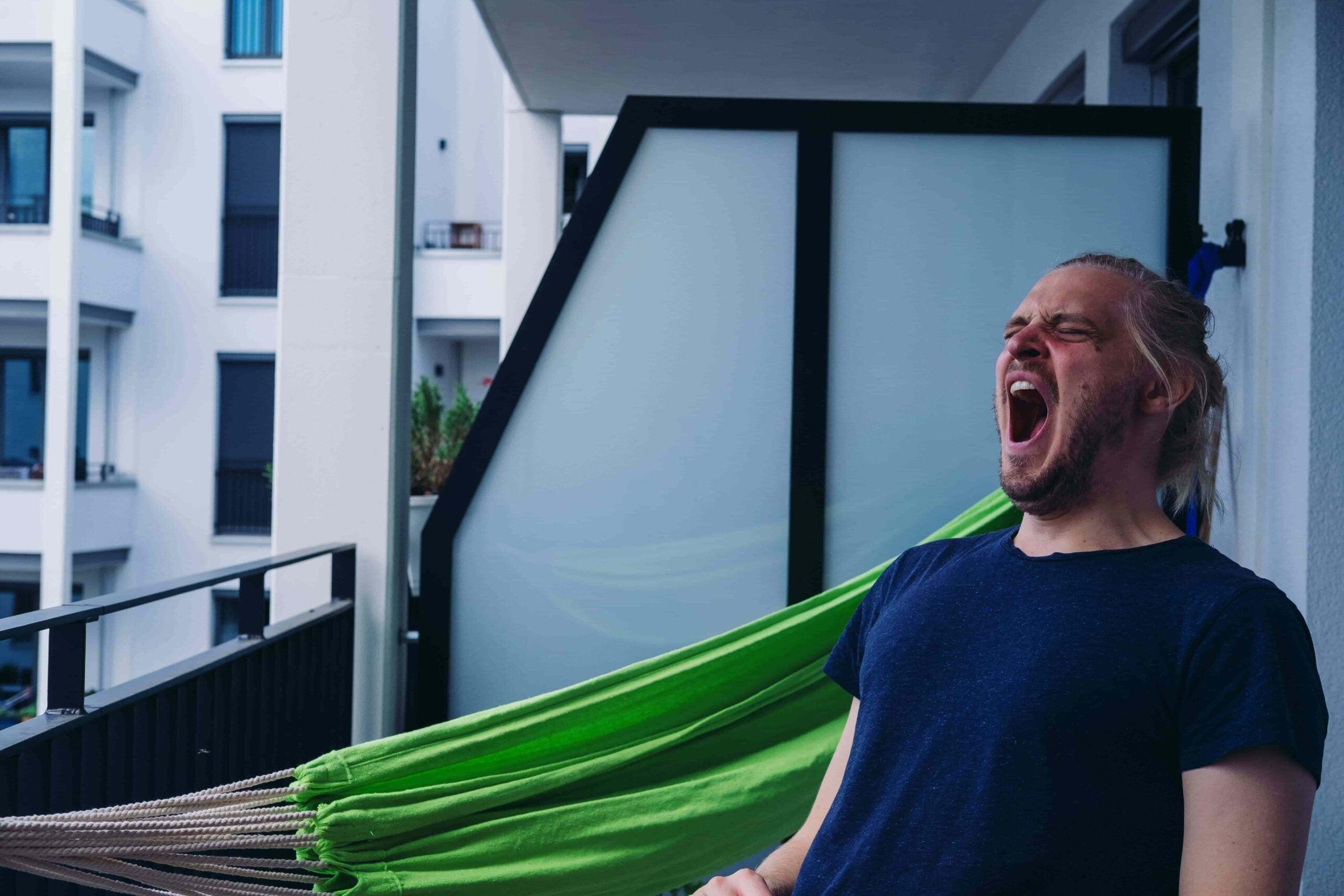 Sleep debt: A man yawning widely while relaxing on a green hammock on a balcony, a common sign of sleepiness or fatigue.
