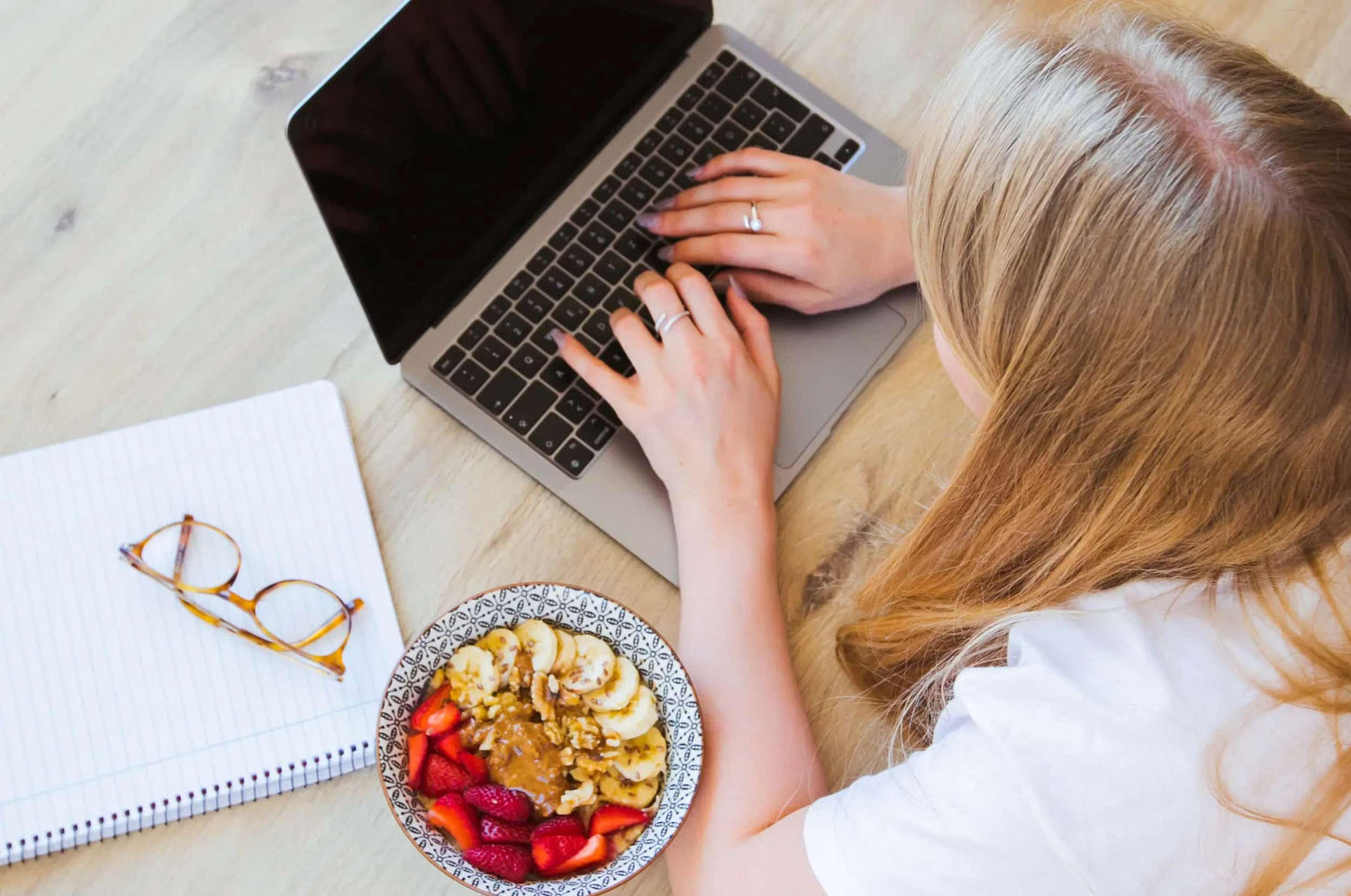 A woman is typing on her laptop with a bowl of fruit and a notebook beside her.