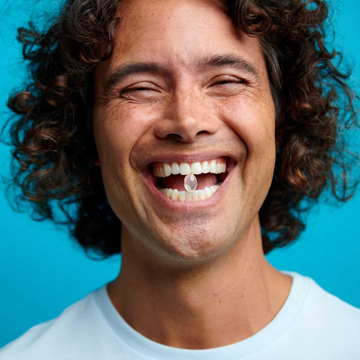 A person with curly hair smiling widely, with a 'Slumber CBN softgel capsule' in their mouth, against a blue backdrop, depicting a sense of well-being and satisfaction associated with the product.